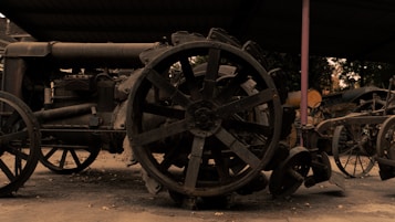 An old, rusty tractor is situated under a metal roof, with large steel wheel spokes and several other vintage farming equipment in the background. The scene seems to be set outdoors with a muted, overcast sky visible through the roof supports.