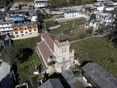 An aerial view of a village with a prominent old church or temple in the center surrounded by greenery and traditional buildings. The area is semi-rural with plots of land, possibly agricultural fields, adjacent to the church. Trees and shrubs add to the rustic charm of the landscape.