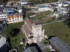 An aerial view of a village with a prominent old church or temple in the center surrounded by greenery and traditional buildings. The area is semi-rural with plots of land, possibly agricultural fields, adjacent to the church. Trees and shrubs add to the rustic charm of the landscape.