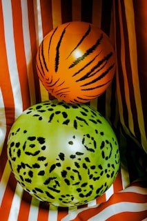 Two balloons with an animal print design are placed against a background of red and white striped fabric. The upper balloon is orange with black tiger-like stripes, while the lower balloon is lime green with a black leopard pattern.