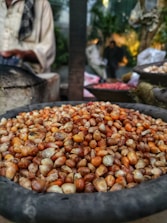 A close-up of a container filled with assorted kernels, predominantly in shades of brown and orange. In the background, a person wearing a light garment, whose face is not visible, stands near some large sacks. The setting appears to be an outdoor market, with foliage visible and a slightly blurred ambiance behind.