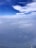Aerial view featuring a vast expanse of cloudy sky with patches of bright white clouds against a deep blue background. Below, a hazy landscape is visible, giving an impression of height and distance.