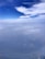 Aerial view featuring a vast expanse of cloudy sky with patches of bright white clouds against a deep blue background. Below, a hazy landscape is visible, giving an impression of height and distance.