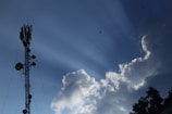 A telecommunications tower stands tall against a clear blue sky, with large fluffy white clouds nearby. Sun rays visibly shine down, creating a dramatic effect. Birds fly in the sky, adding to the serene atmosphere.