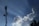 A telecommunications tower stands tall against a clear blue sky, with large fluffy white clouds nearby. Sun rays visibly shine down, creating a dramatic effect. Birds fly in the sky, adding to the serene atmosphere.