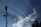 A telecommunications tower stands tall against a clear blue sky, with large fluffy white clouds nearby. Sun rays visibly shine down, creating a dramatic effect. Birds fly in the sky, adding to the serene atmosphere.