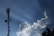 A telecommunications tower stands tall against a clear blue sky, with large fluffy white clouds nearby. Sun rays visibly shine down, creating a dramatic effect. Birds fly in the sky, adding to the serene atmosphere.