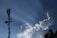 A telecommunications tower stands tall against a clear blue sky, with large fluffy white clouds nearby. Sun rays visibly shine down, creating a dramatic effect. Birds fly in the sky, adding to the serene atmosphere.
