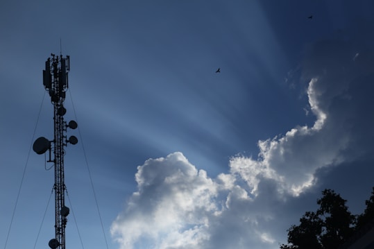 A telecommunications tower stands tall against a clear blue sky, with large fluffy white clouds nearby. Sun rays visibly shine down, creating a dramatic effect. Birds fly in the sky, adding to the serene atmosphere.