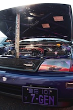 A customized car engine is visible with the hood open, featuring a prominently displayed rod with the word 'Corolla' written vertically. The engine area is clean and includes various components with red and black accents. A 'Touring Wagon' badge is on the grille. The vehicle has a blue exterior with a distinctive Japanese license plate reading '7-GEN'.