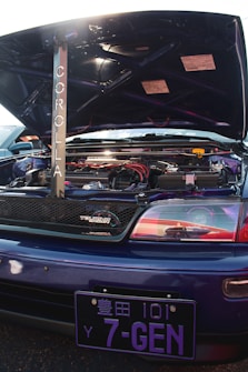A customized car engine is visible with the hood open, featuring a prominently displayed rod with the word 'Corolla' written vertically. The engine area is clean and includes various components with red and black accents. A 'Touring Wagon' badge is on the grille. The vehicle has a blue exterior with a distinctive Japanese license plate reading '7-GEN'.