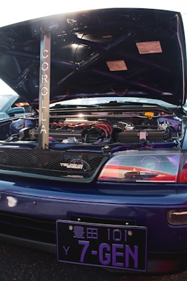 A customized car engine is visible with the hood open, featuring a prominently displayed rod with the word 'Corolla' written vertically. The engine area is clean and includes various components with red and black accents. A 'Touring Wagon' badge is on the grille. The vehicle has a blue exterior with a distinctive Japanese license plate reading '7-GEN'.