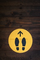 A wooden floor background features a circular yellow sign with two black footprints and an upward-pointing arrow.
