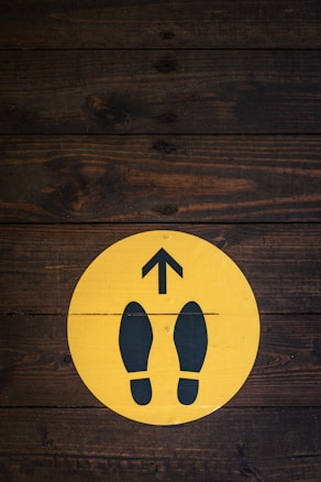 A wooden floor background features a circular yellow sign with two black footprints and an upward-pointing arrow.