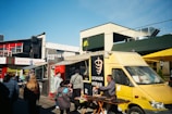 A bustling street scene with a yellow food truck offering doner kebab parked next to a sidewalk. Several people are gathered around the truck and others walk by, including a man cooking on a portable stove. Surrounding buildings include a pub and various shops. A clear blue sky provides background, while a yellow umbrella offers shade over some tables.