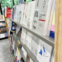 A bookstore or library shelf filled with various books is on display. The books have different sizes, colors, and covers. Some titles are visible, and there are multiple rows of books organized neatly. The background shows a large window with greenery visible outside.