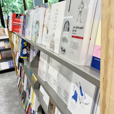 A bookstore or library shelf filled with various books is on display. The books have different sizes, colors, and covers. Some titles are visible, and there are multiple rows of books organized neatly. The background shows a large window with greenery visible outside.