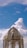 A person is standing on scaffolding while working on the restoration or painting of a dome-like structure. The sky is bright and clear with some scattered clouds. The structure appears weathered with peeling paint, and there are decorative elements and round finials around the top.
