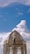 A person is standing on scaffolding while working on the restoration or painting of a dome-like structure. The sky is bright and clear with some scattered clouds. The structure appears weathered with peeling paint, and there are decorative elements and round finials around the top.