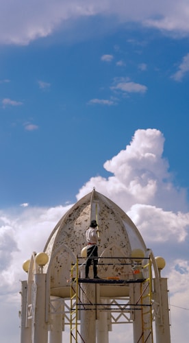 A person is standing on scaffolding while working on the restoration or painting of a dome-like structure. The sky is bright and clear with some scattered clouds. The structure appears weathered with peeling paint, and there are decorative elements and round finials around the top.