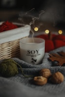 A soy wax candle labeled 'white tea' is nestled among cozy autumn objects. There is a wooden basket with red yarn and blurred bokeh lights in the background. The scene includes a gray fabric, walnuts, a green ball of yarn, and a brown leaf, creating a warm, rustic atmosphere.