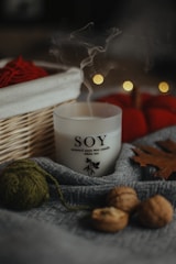 A soy wax candle labeled 'white tea' is nestled among cozy autumn objects. There is a wooden basket with red yarn and blurred bokeh lights in the background. The scene includes a gray fabric, walnuts, a green ball of yarn, and a brown leaf, creating a warm, rustic atmosphere.