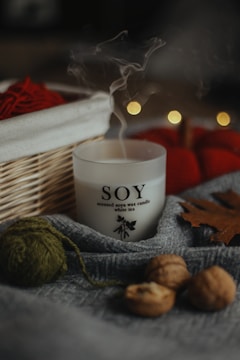 A soy wax candle labeled 'white tea' is nestled among cozy autumn objects. There is a wooden basket with red yarn and blurred bokeh lights in the background. The scene includes a gray fabric, walnuts, a green ball of yarn, and a brown leaf, creating a warm, rustic atmosphere.