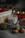 A soy wax candle labeled 'white tea' is nestled among cozy autumn objects. There is a wooden basket with red yarn and blurred bokeh lights in the background. The scene includes a gray fabric, walnuts, a green ball of yarn, and a brown leaf, creating a warm, rustic atmosphere.