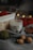 A soy wax candle labeled 'white tea' is nestled among cozy autumn objects. There is a wooden basket with red yarn and blurred bokeh lights in the background. The scene includes a gray fabric, walnuts, a green ball of yarn, and a brown leaf, creating a warm, rustic atmosphere.