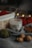 A soy wax candle labeled 'white tea' is nestled among cozy autumn objects. There is a wooden basket with red yarn and blurred bokeh lights in the background. The scene includes a gray fabric, walnuts, a green ball of yarn, and a brown leaf, creating a warm, rustic atmosphere.