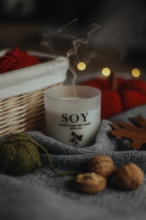 A soy wax candle labeled 'white tea' is nestled among cozy autumn objects. There is a wooden basket with red yarn and blurred bokeh lights in the background. The scene includes a gray fabric, walnuts, a green ball of yarn, and a brown leaf, creating a warm, rustic atmosphere.