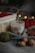 A soy wax candle labeled 'white tea' is nestled among cozy autumn objects. There is a wooden basket with red yarn and blurred bokeh lights in the background. The scene includes a gray fabric, walnuts, a green ball of yarn, and a brown leaf, creating a warm, rustic atmosphere.