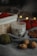 A soy wax candle labeled 'white tea' is nestled among cozy autumn objects. There is a wooden basket with red yarn and blurred bokeh lights in the background. The scene includes a gray fabric, walnuts, a green ball of yarn, and a brown leaf, creating a warm, rustic atmosphere.