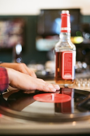 A person gently places hands on a spinning vinyl record on a turntable. A liquor bottle with a red label is positioned in the background on a table, creating a relaxed, vintage ambiance.