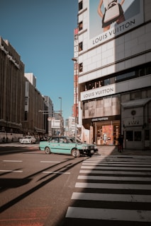 A city street scene featuring a prominent Louis Vuitton store on a building facade. A turquoise taxi is crossing a pedestrian crosswalk. The urban setting includes tall buildings and billboards under a clear blue sky.