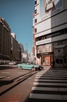 A city street scene featuring a prominent Louis Vuitton store on a building facade. A turquoise taxi is crossing a pedestrian crosswalk. The urban setting includes tall buildings and billboards under a clear blue sky.