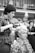 A woman in a checkered shirt styles another woman's blonde hair. The seated woman wears a cape with a floral pattern, and her expression appears thoughtful. In the background, several people are visible in what seems like a public space.