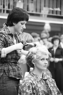 A woman in a checkered shirt styles another woman's blonde hair. The seated woman wears a cape with a floral pattern, and her expression appears thoughtful. In the background, several people are visible in what seems like a public space.