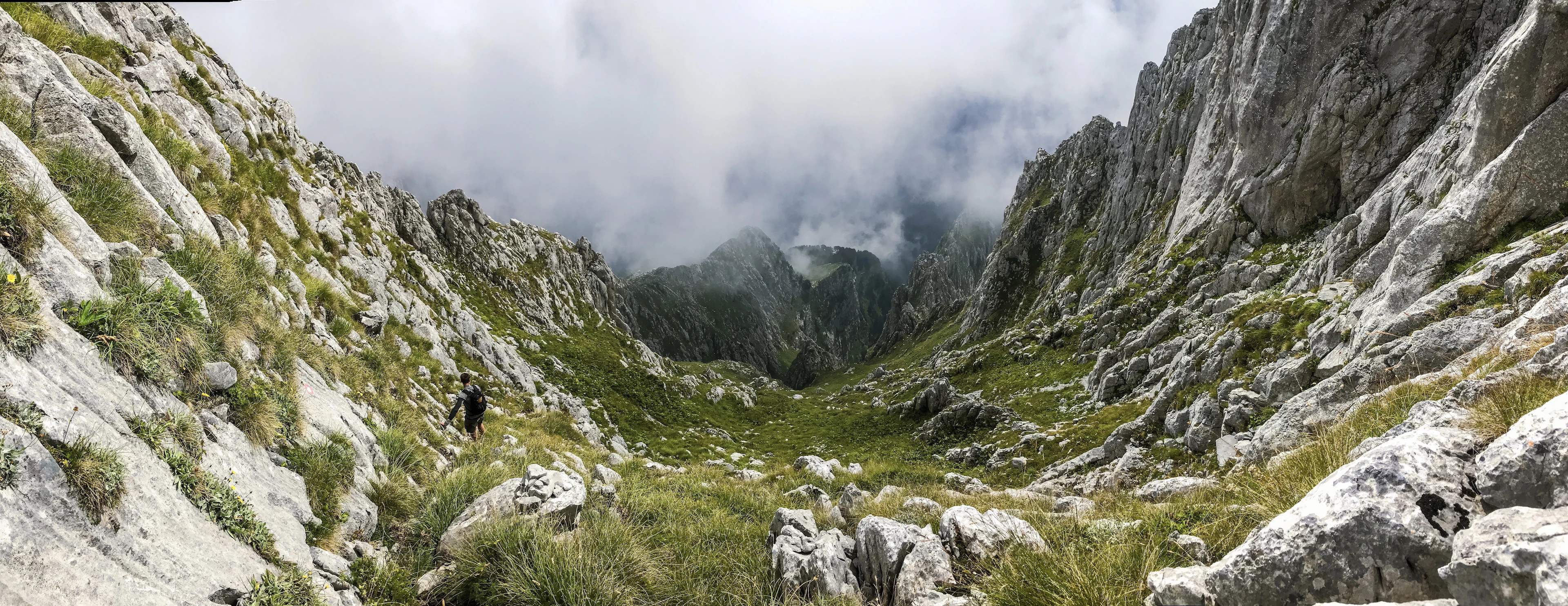 A hiker climbing a steep, grassy limestone valley in Prokletije National Park, with the peaks wrapped in cloud