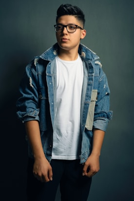 A person wearing a white t-shirt and a distressed denim jacket stands against a dark gray background, looking slightly off-camera. The jacket has a few rips and patches, adding a casual, trendy look to the outfit. The individual is also wearing black-rimmed glasses and has short, styled hair.