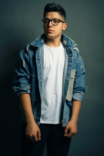 A person wearing a white t-shirt and a distressed denim jacket stands against a dark gray background, looking slightly off-camera. The jacket has a few rips and patches, adding a casual, trendy look to the outfit. The individual is also wearing black-rimmed glasses and has short, styled hair.