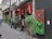 A narrow street with a vibrant café featuring a colorful exterior with plants and artwork. The artwork includes abstract figures in bright colors, and there are small flags above the entrance. People are casually standing and walking.