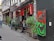 A narrow street with a vibrant café featuring a colorful exterior with plants and artwork. The artwork includes abstract figures in bright colors, and there are small flags above the entrance. People are casually standing and walking.