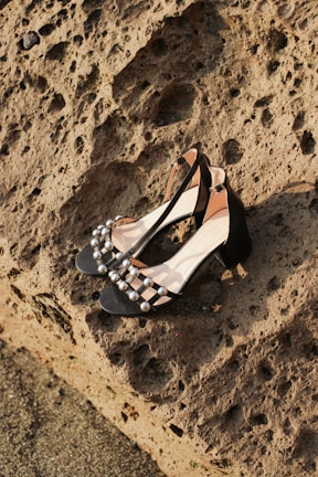 A pair of black high-heeled shoes adorned with metallic studs, placed on a rugged, textured stone surface. The shoes feature a smooth, shiny finish with a classic design, contrasting with the raw, uneven background.