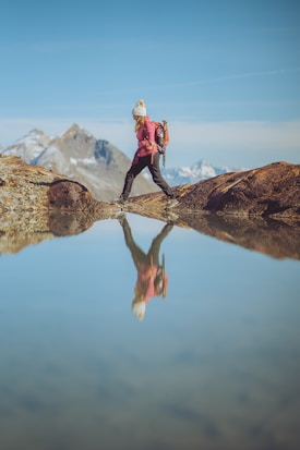 A person wearing a pink jacket, black pants, and a white hat is hiking across a rocky path beside a reflective body of water. The landscape includes distant mountains under a clear blue sky, and the hiker