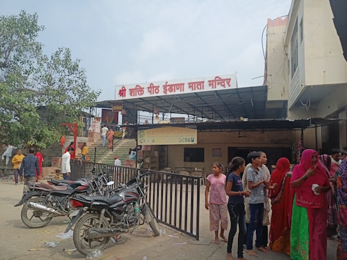 A busy outdoor area in front of a temple, which features a sign with text in a regional script. Several people, including women and children, stand near metal railings in colorful traditional clothing. Motorcycles are parked nearby, and a large tree is visible in the background. The temple entrance is elevated with stairs leading up to it.