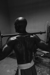 A muscular man is lifting a barbell with weights in a dimly lit room. The focus is on his back muscles, glistening from exertion. The floor appears to be tiled, and some gym equipment is visible in the background.