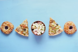 A minimalistic composition featuring a symmetrical arrangement of pastries, pizza slices, and a bowl of popcorn on a light blue background. The pastries have a glossy appearance with a purple filling at the center, while the pizza slices are topped with vegetables like olives, mushrooms, and peppers. The bowl in the middle contains colorful popcorn.