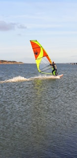 A person is windsurfing on a large body of water. The windsurfer is moving at a swift pace, creating a trail of splashes behind. The sail is brightly colored with yellow, orange, and green sections, and a distinctive black design. The background features a distant shoreline with trees and a clear blue sky.