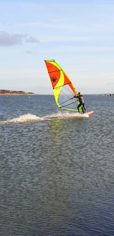 A person is windsurfing on a large body of water. The windsurfer is moving at a swift pace, creating a trail of splashes behind. The sail is brightly colored with yellow, orange, and green sections, and a distinctive black design. The background features a distant shoreline with trees and a clear blue sky.